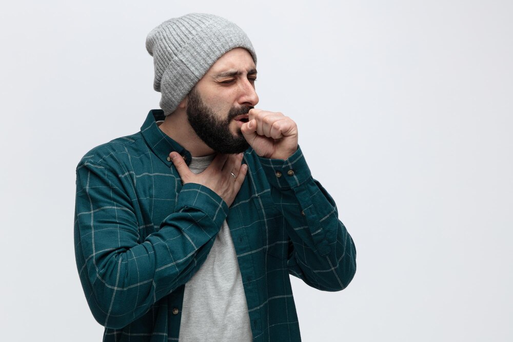 Unhealthy young man wearing winter hat keeping hand on chest and fist in front of mouth coughing with closed eyes isolated on white background with copy space